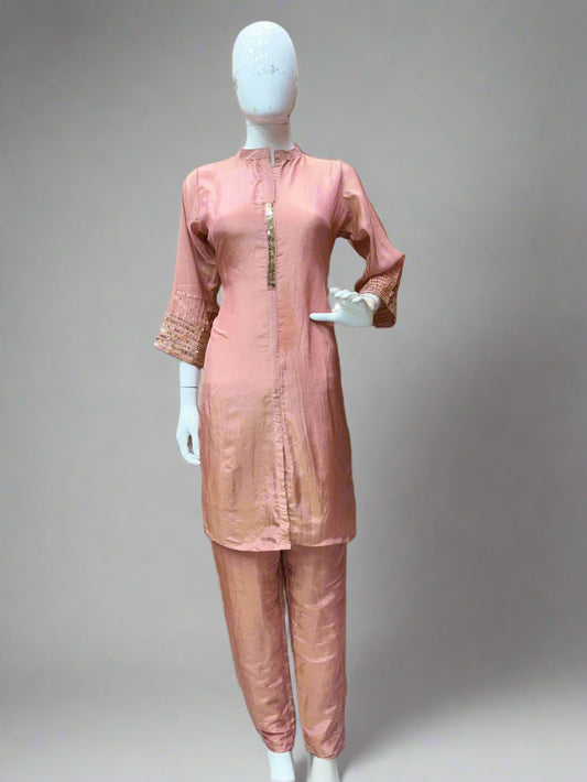 Mannequin wearing a pink traditional outfit with a beige wall and colorful clothes in the background.