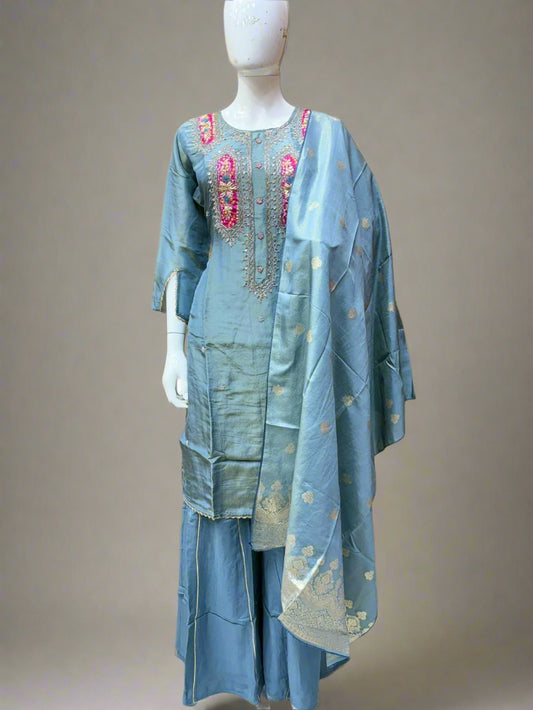 Mannequin wearing a light blue traditional outfit with embroidery in a room setting.