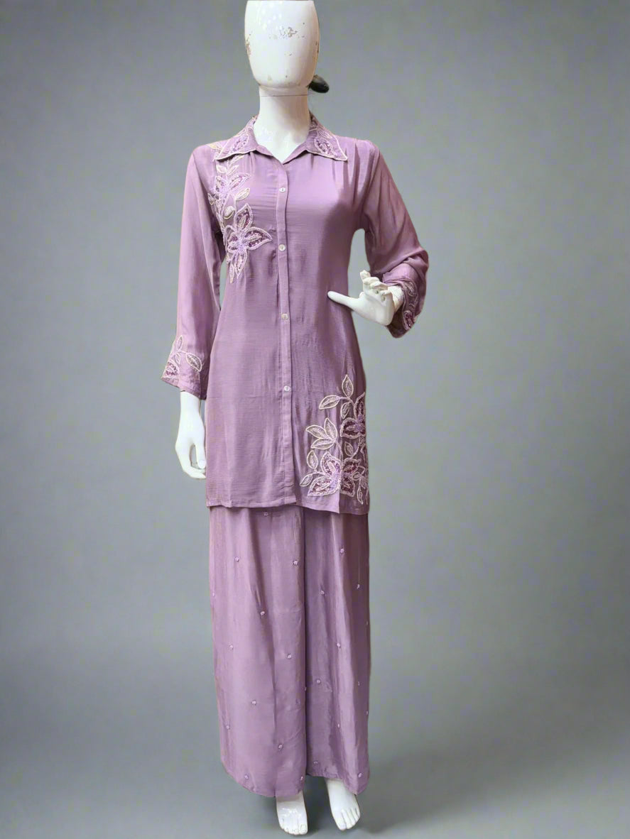 Mannequin wearing a mauve traditional outfit with embroidery, standing against a beige wall with colorful garments hanging on a rack.